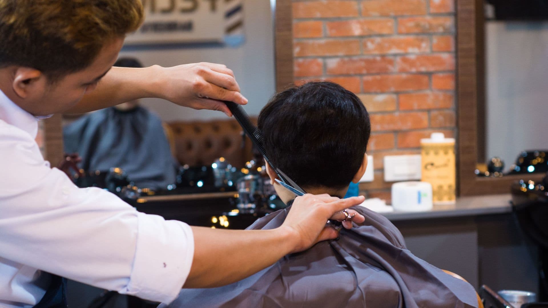 Barbeiro a cortar o cabelo de um cliente numa barbearia com parede de tijolo