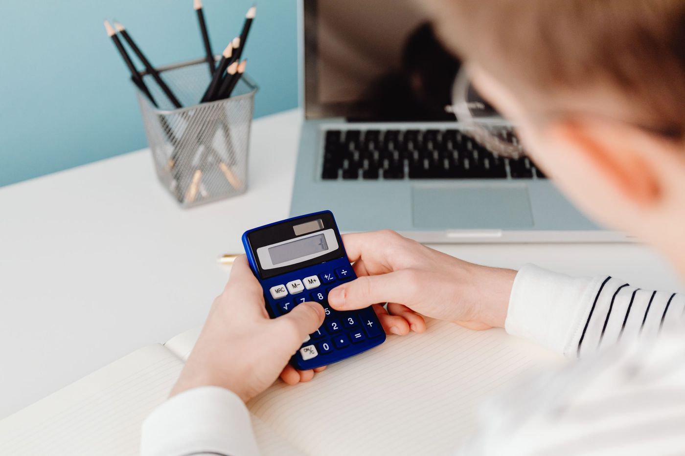 Mãos a usar uma calculadora azul numa secretária com portátil